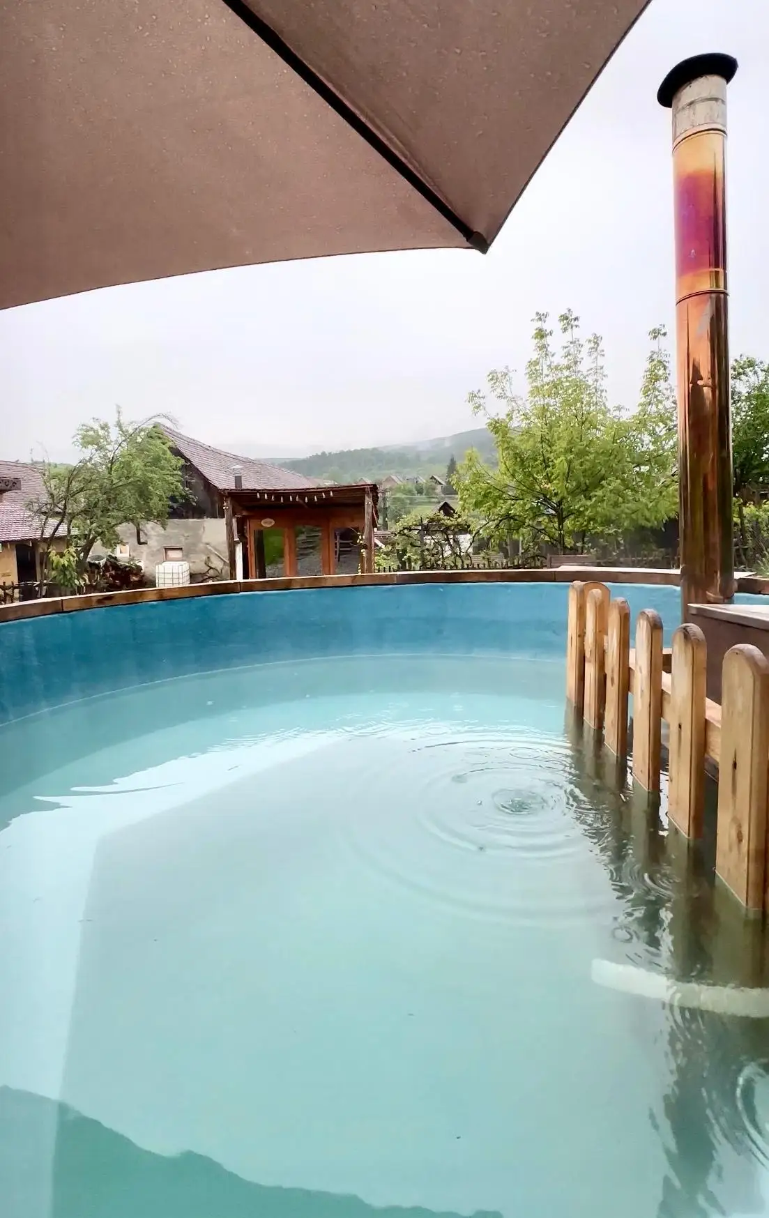 The wood-fired hot tub on the deck with its copper chimney, the village rooftops and green hills beyond on a rainy day