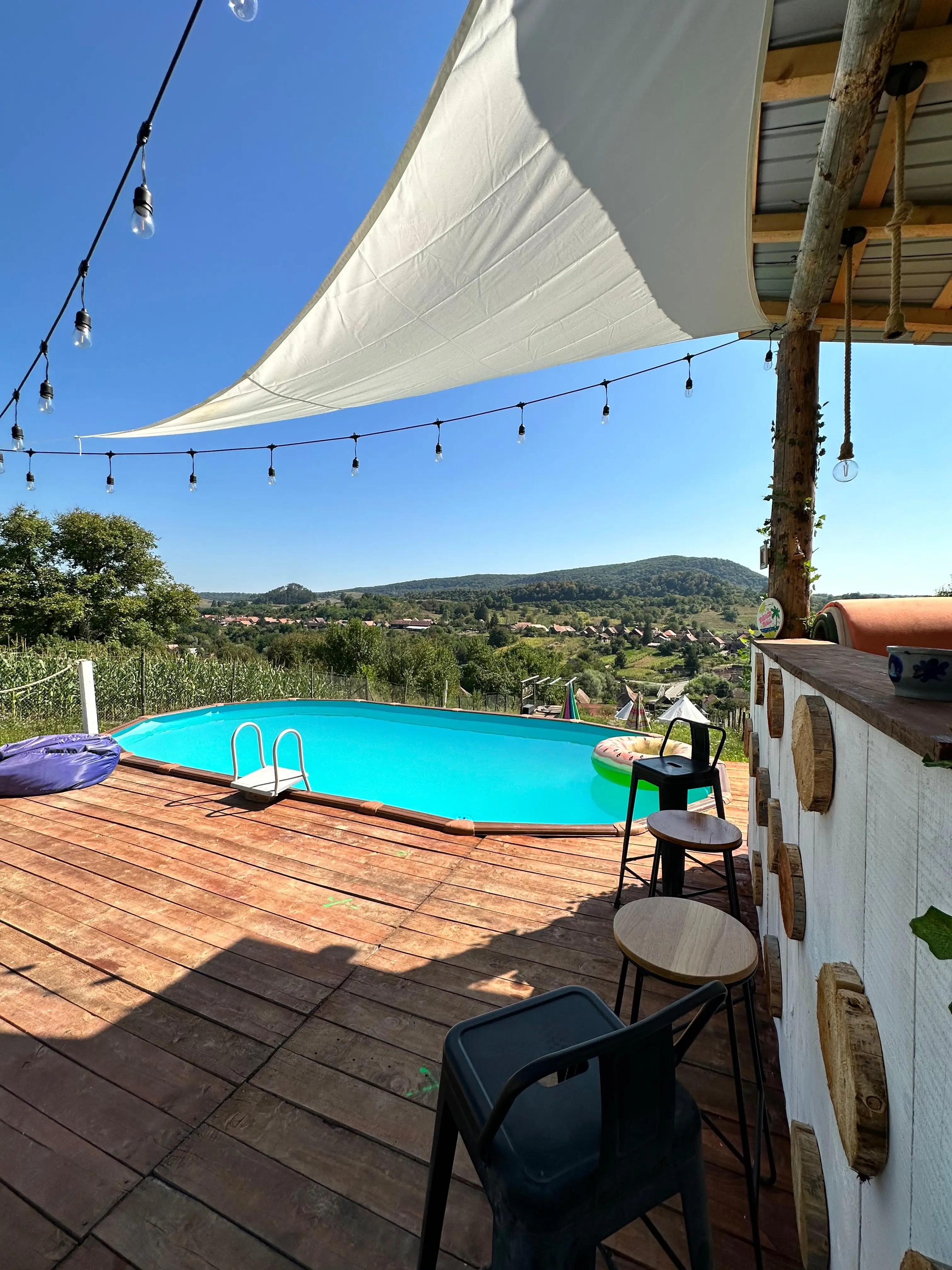 The pool on its wooden deck under a white sail shade, a bar with stools alongside, looking out over the village of Saschiz and the hills beyond